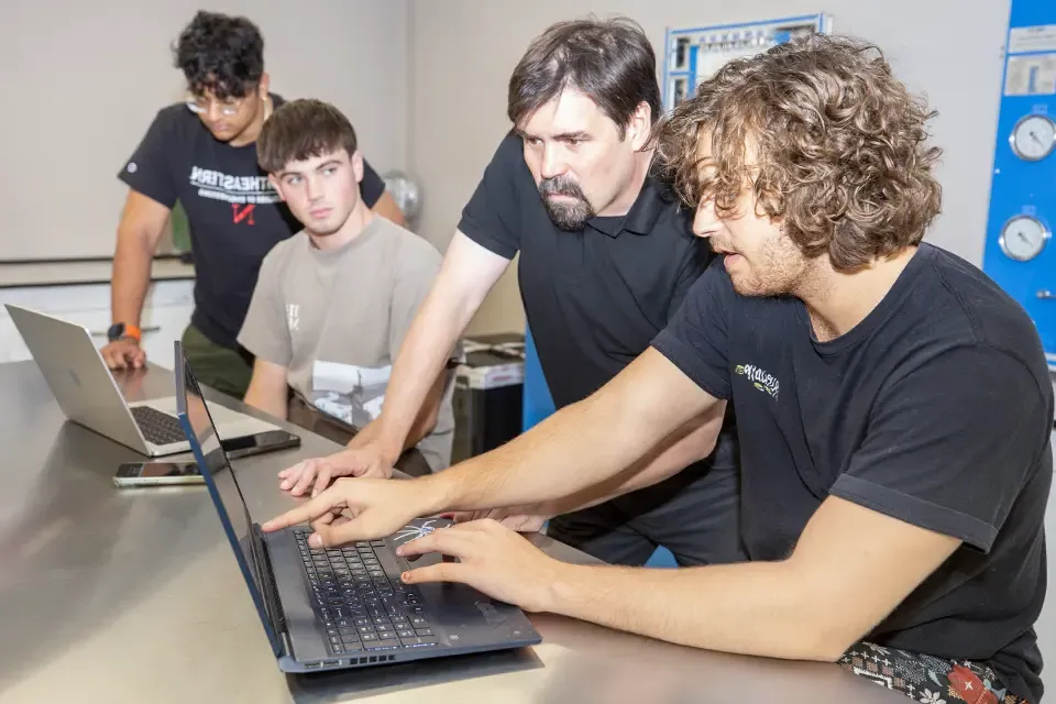 Two students and an instructor examine a computer screen while having a discussion.